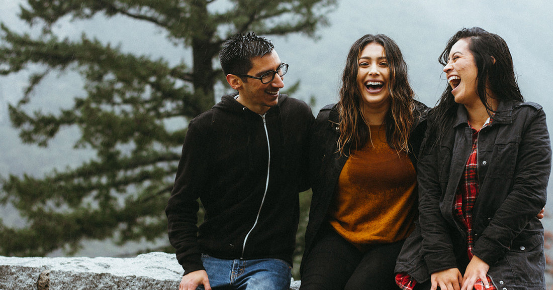 three smiling people sitting on ledge in from of large evergreen tree