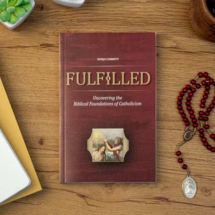 A tabletop lifestyle shot of the Catholic book, Fulfilled: Uncovering the Biblical Foundations of Catholicism by Sonja Corbitt, sitting on wooden table next to a red rosary. The red cover features the Baptism of Jesus at the Jordan.