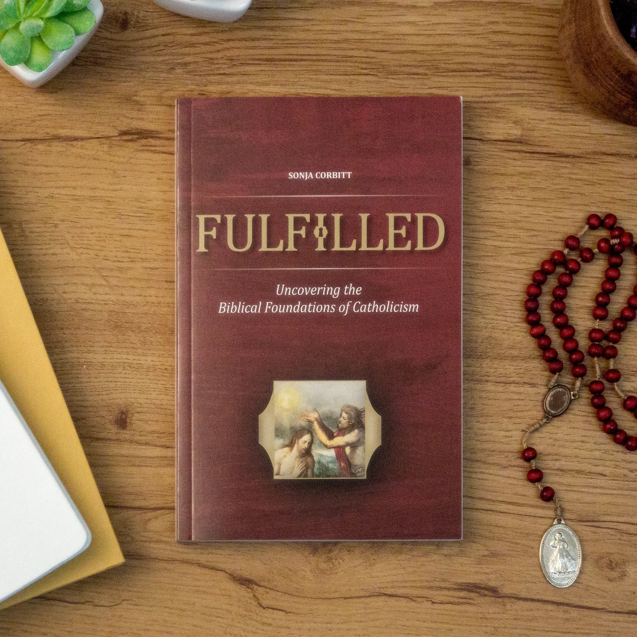 A tabletop lifestyle shot of the Catholic book, Fulfilled: Uncovering the Biblical Foundations of Catholicism by Sonja Corbitt, sitting on wooden table next to a red rosary. The red cover features the Baptism of Jesus at the Jordan.