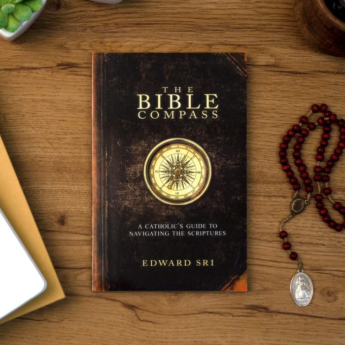 A tabletop lifestyle shot of the catholic book, The Bible Compass: A Catholic's Guide to Navigating the Scriptures by Edward Sri published by Ascension, sitting on a wooden table. The brown cover features a gold compass.