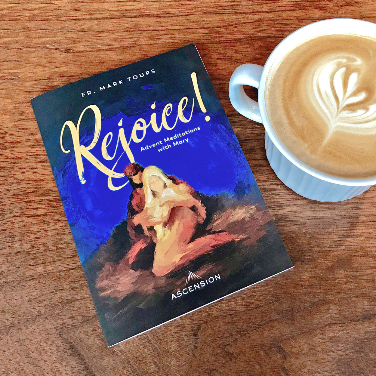 A copy of the Catholic journal, Rejoice! Advent Meditations with Mary by Fr. Mark Toups and Ascension on a wooden table next to a cappuccino.